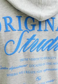 Grijze hoodie met een groot blauw grafisch tekstontwerp, met zinnen in verschillende lettertypen en -groottes, waaronder "ORIGINAL" en "STUDIO."