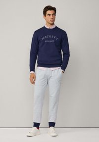 Marine sweatshirt med teksten "HACKETT LONDON", grå sweatpants med logo og hvide sneakers. Stribet skjortekrave er synlig under sweatshirten.