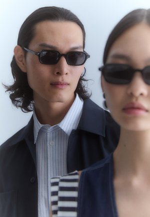 Occhiali da sole neri con montatura angolare indossati da un uomo e una donna. L'uomo ha una camicia a righe blu e bianche sotto una giacca scura; la donna indossa un top scuro e a fantasia.