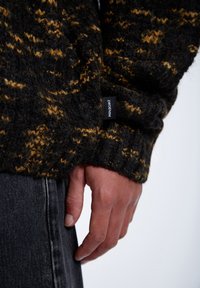 Czarny i żółty dziergany sweter z fakturą i boczną metką z napisem "DR.PDENIM." Ręka spoczywa na ciemnych spodniach dżinsowych.