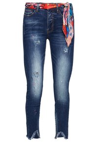 Tunika blå denimjeans med slitna detaljer, fransade fållar och ett färgglatt blommigt scarvesbälte. Har fickor och en klassisk femficksdesign.