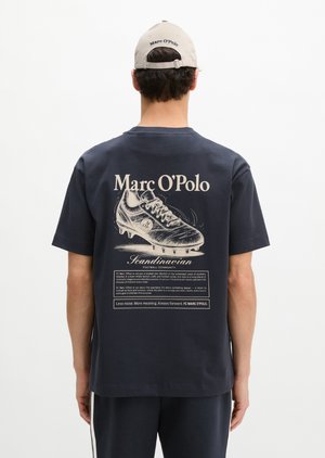 Homme portant un t-shirt bleu marine avec un grand motif de crampon de football et le texte « Marc O’Polo », assorti d'une casquette beige Marc O’Polo, de dos.