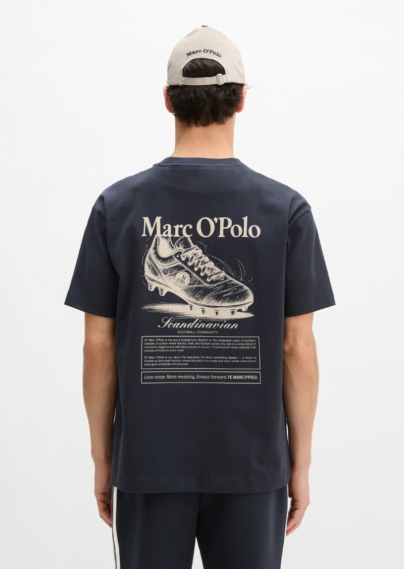 Άνδρας με μπλε σκούρο t-shirt με μεγάλο γραφικό ενός ποδοσφαιρικού παπουτσιού και το κείμενο «Marc O'Polo», συνδυασμένο με μπεζ καπέλο Marc O'Polo, με την πλάτη προς τα έξω.