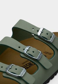 Κοντινή λήψη πράσινων λουριών δερμάτινου σανδαλιού με ασημένιες μεταλλικές πόρπες και ανοιχτό καφέ πέλμα.