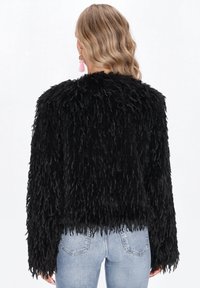 Frau mit welligem blonden Haar, die eine strukturierte schwarze Jacke mit langen Ärmeln und hellblauen Jeans trägt, von hinten gezeigt.