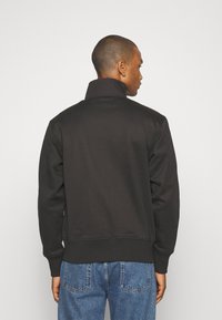Felpa nera con colletto alto, caratterizzata da polsini e orlo a coste. Tessuto liscio, mostrata dal retro, abbinata a jeans blu.