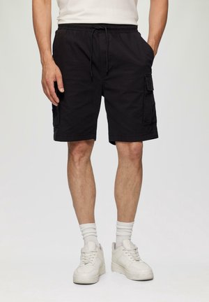 Mann trägt schwarze Cargo-Shorts mit Kordelzug in der Taille, weiße Socken und weiße Sneakers, steht mit den Händen in den Taschen.
