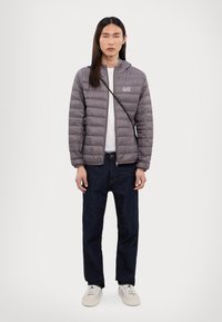 Grå puffjakke med horisontalt quiltet tekstur, glidelås foran og hette. Kombinert med mørke denimjeans og hvite sneakers. Enkelt merkevarelogo på jakken.