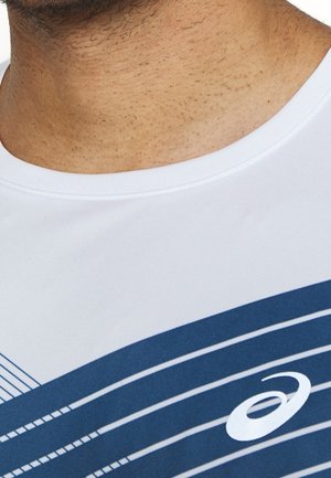 Zbliżenie na szyję i górną część klatki piersiowej osoby noszącej białą koszulę z niebieskimi ukośnymi liniami i białym logo Asics.