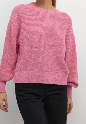 Femme portant un pull en maille rose ample avec des poignets et un ourlet côtelés, associé à un pantalon noir sur un fond uni.