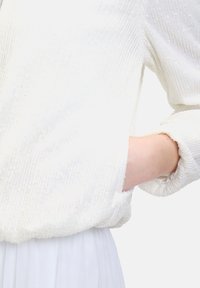 Weißes, strukturiertes Oberteil mit geripptem Muster und subtilen Pailletten. Es hat eine entspannte Passform mit umgeschlagenen Ärmeln und einer Tasche auf der linken Seite.