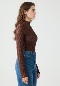 Bruin getextureerde lange mouw top met een hoge kraag, gecombineerd met hooggeknoopte blauwe spijkerbroek. Zichtbare stofdetails op de mouwen.