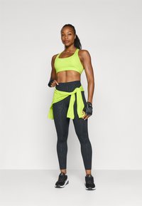 Sujeción deportiva verde neón con leggins negros de cintura alta que presentan un patrón texturizado, combinados con una camiseta verde neón atada y zapatillas deportivas negras.