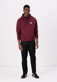 Sweat-shirt à capuche bordeaux avec poche avant et logo, associé à un pantalon noir et des baskets noires avec des rayures blanches sur les côtés.