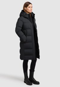 Čierna vatovaná bunda s kapucňou, so štepovaným dizajnom, hladkou textúrou, dvoma bočnými vreckami na zips a siluetou do dĺžky kolien.