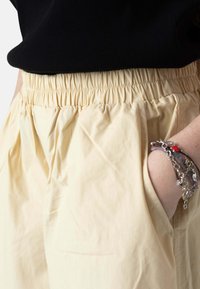 Persona con pantalones beige de cintura elástica con la mano derecha en el bolsillo, camiseta negra y varias pulseras de cadena en la muñeca.