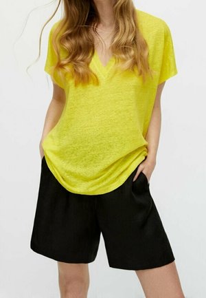 Femme portant un haut ample jaune à col en V et manches courtes, légèrement rentré dans un short noir longueur genou, les mains dans les poches.