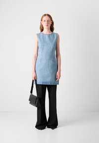 Τζιν φόρεμα χωρίς μανίκια σε ανοιχτό μπλε με στενή εφαρμογή, συνδυασμένο με φαρδιά μαύρα παντελόνια. Το μοντέλο κρατά μια μικρή μαύρη τσάντα χιαστί.