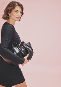 Czarne skórzane torby trzymane przy dopasowanej czarnej sukience, o gładkiej teksturze i różnych kształtach. Delikatnie różowe tło podkreśla kontrast.