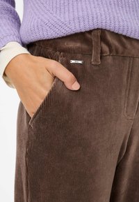 Bruine corduroy broek met verticale ribbels, voorzien van een zijzak en een zilverkleurig metalen label met "Brazz." Draag het met een licht lila trui.