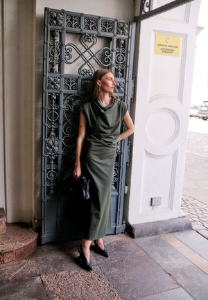 Femme en robe verte sans manches et talons noirs se tient contre une porte en métal ornée près de l'entrée avec un panneau de l'ambassade du Monténégro.