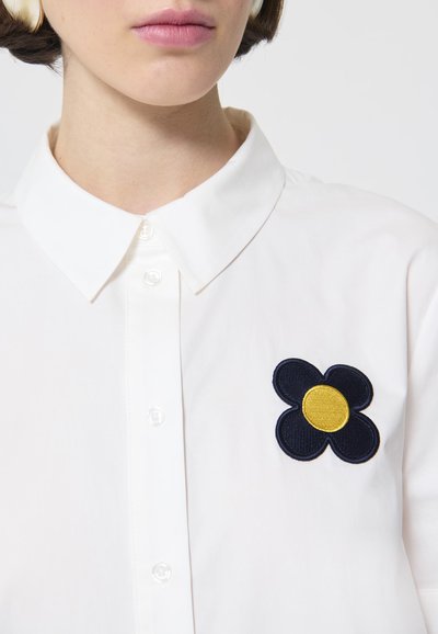 Witte blouse met een puntige kraag en een geborduurde marineblauwe bloem met een gele kern aan de linkerkant. Gladde stofstructuur.