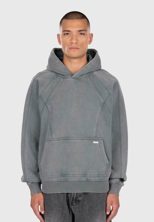 Uomo che indossa un ampio hoodie grigio spento con tasca a marsupio frontale e polsini a costine, abbinato a jeans neri sbiaditi.