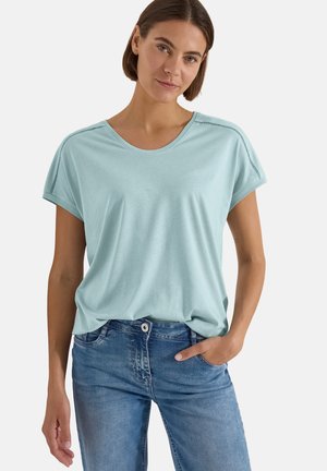 Donna con capelli corti castani che indossa una maglietta azzurro chiaro a maniche corte e jeans blu, in piedi con una mano in tasca.