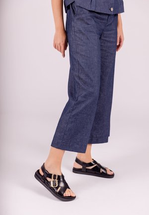 Sandalias de cuero negro con acentos de doble hebilla, combinadas con pantalones cortos de corte holgado en azul marino, hechos de tela texturizada.