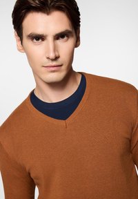 Brązowy sweter w serek wykonany z delikatnego materiału, nałożony na granatową koszulę. Tkanina ma gładką fakturę z prążkowanymi akcentami.
