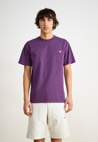 Camiseta de algodón morada con mangas cortas, con un pequeño logo rojo y amarillo en el pecho, combinada con pantalones cortos gris claro.