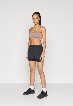 Světle hnědá sportovní podprsenka s tenkými ramínky, doplněná černými šortkami a černými teniskami. Bílá členková ponožka zvýrazňuje outfit.