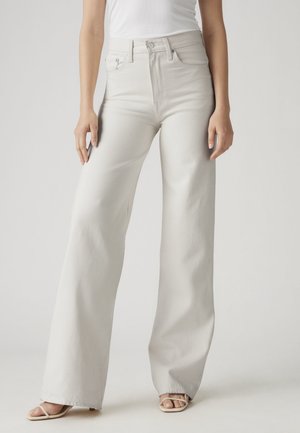 Femme portant un jean blanc taille haute à jambes larges, associé à des talons ouverts et à lanières ainsi qu'un haut blanc sans manches, debout devant un fond uni.