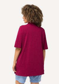 Bordová tunika s krátkými rukávy, má rovný lem a boční rozparky, vyrobena z měkké bavlny, nošena přes světle modré šortky.