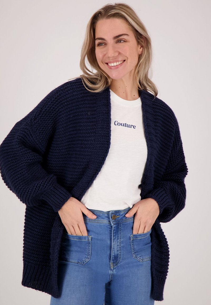 Glimlachende vrouw met blond haar in een marineblauwe grof gebreide cardigan, een witte t-shirt met de tekst "Couture" en blauwe high-waisted jeans met haar handen in de zakken.