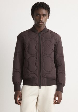 Young man with dark skin and short curly hair wearing a dark brown quilted zip-up jacket and light beige pants against a plain background.
