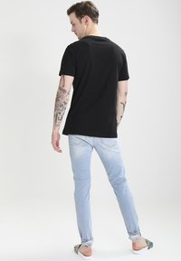 Camiseta negra de algodón con cuello redondo, combinada con jeans de mezclilla azul claro. Tatuajes notables visibles en los brazos, modelo de espaldas a la cámara.