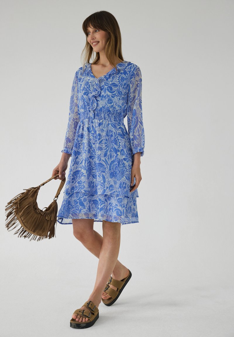 Femme portant une robe bleue à fleurs mi-longue, tenant un sac à main marron à franges et portant des sandales à boucles couleur camel sur fond blanc.