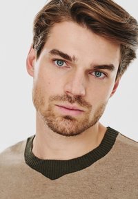 Brązowy sweter dżersejowy z ciemnozielonym prążkowanym kołnierzem, o gładkiej fakturze. Zbliżenie na twarz męskiego modela z jasnobrązowymi włosami i niebieskimi oczami.