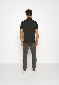 Man står vänd mot en vit vägg, iklädd en svart kortärmad tröja, mörkgrå jeans och vita sneakers.