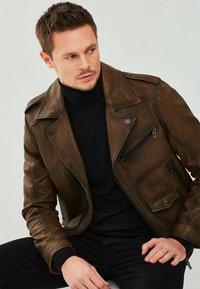 Veste de motard en cuir marron avec épaulettes, poches zippées et finition texturée, portée par-dessus un col roulé noir.