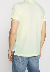 Svetle žlutá polo tričko s krátkým rukávem a límečkem, vyrobené z měkké tkaniny, s bočními rozparky a rovnými lemy, nošeno s modrými džínami.