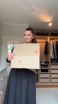 Kartonasti škatla z logotipom Zalando v oranžni barvi, postavljena pred svetlo obarvane stene sobe, z oblačili, ki so vidna v odprtem omari.