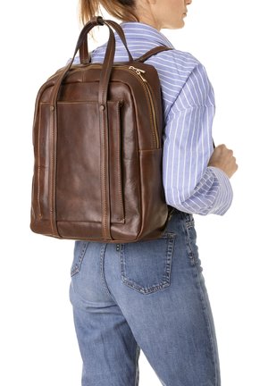 Vrouw draagt een blauw gestreept shirt en spijkerbroek en heeft een bruine leren rugzak met dubbele handgrepen en ritscompartimenten bij zich.