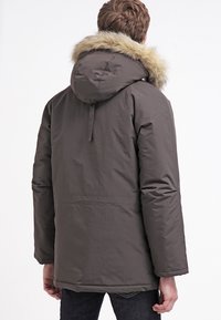 Parka giacca di colore marrone scuro con cappuccio e bordo in pelliccia sintetica, maniche lunghe e vestibilità leggermente ampia, realizzata in un tessuto morbido.