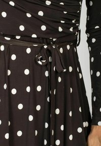 Czarna sukienka w białe kropki, z kopertowym krojem i wiązaniem w talii. Materiał jest gładki, o miękkiej teksturze.