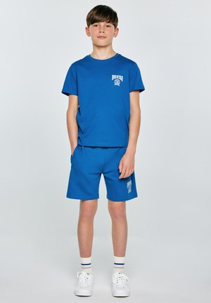 Chłopiec stojący, ubrany w niebieski T-shirt i pasujące do niego spodenki, białe trampki i skarpetki na jednolitym tle.