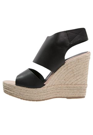 Sandalias de cuña negras de cuero con una banda ancha sobre el pie, plataforma con textura de cuerda y una altura de aproximadamente 10 cm.