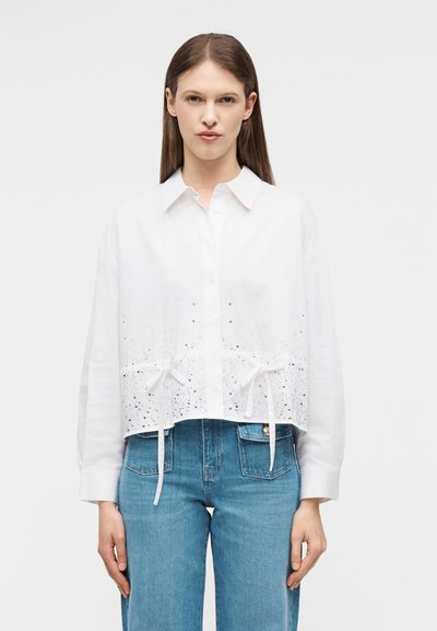 Femme portant une chemise blanche à manches longues boutonnée avec un motif tacheté et des liens à la taille, associée à un jean bleu taille haute.