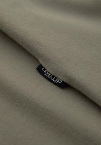 Tejido verde oliva con una textura suave, doblado para revelar una etiqueta negra con "LEVEL UP" en letras blancas.
